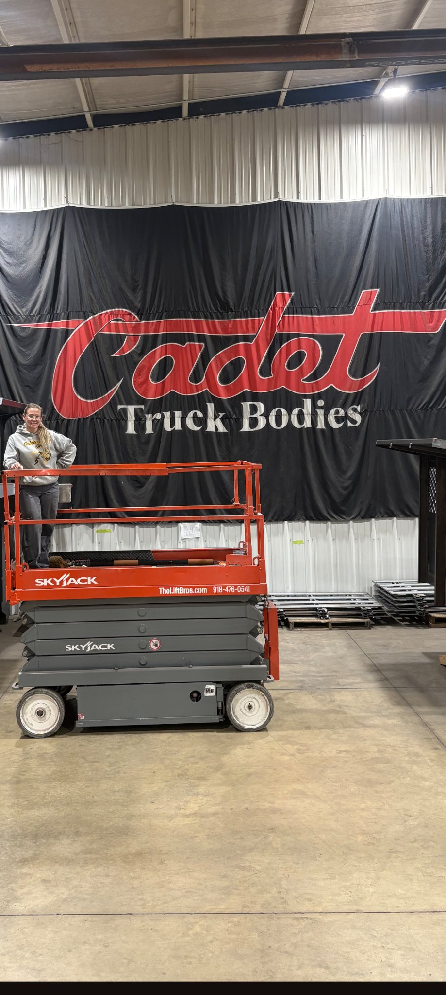 Skyjack scissor lift from The Lift Bros inside the Cadet Truck Bodies shop in Chouteau, Oklahoma, parked in front of the red and black Cadet Truck Bodies banner