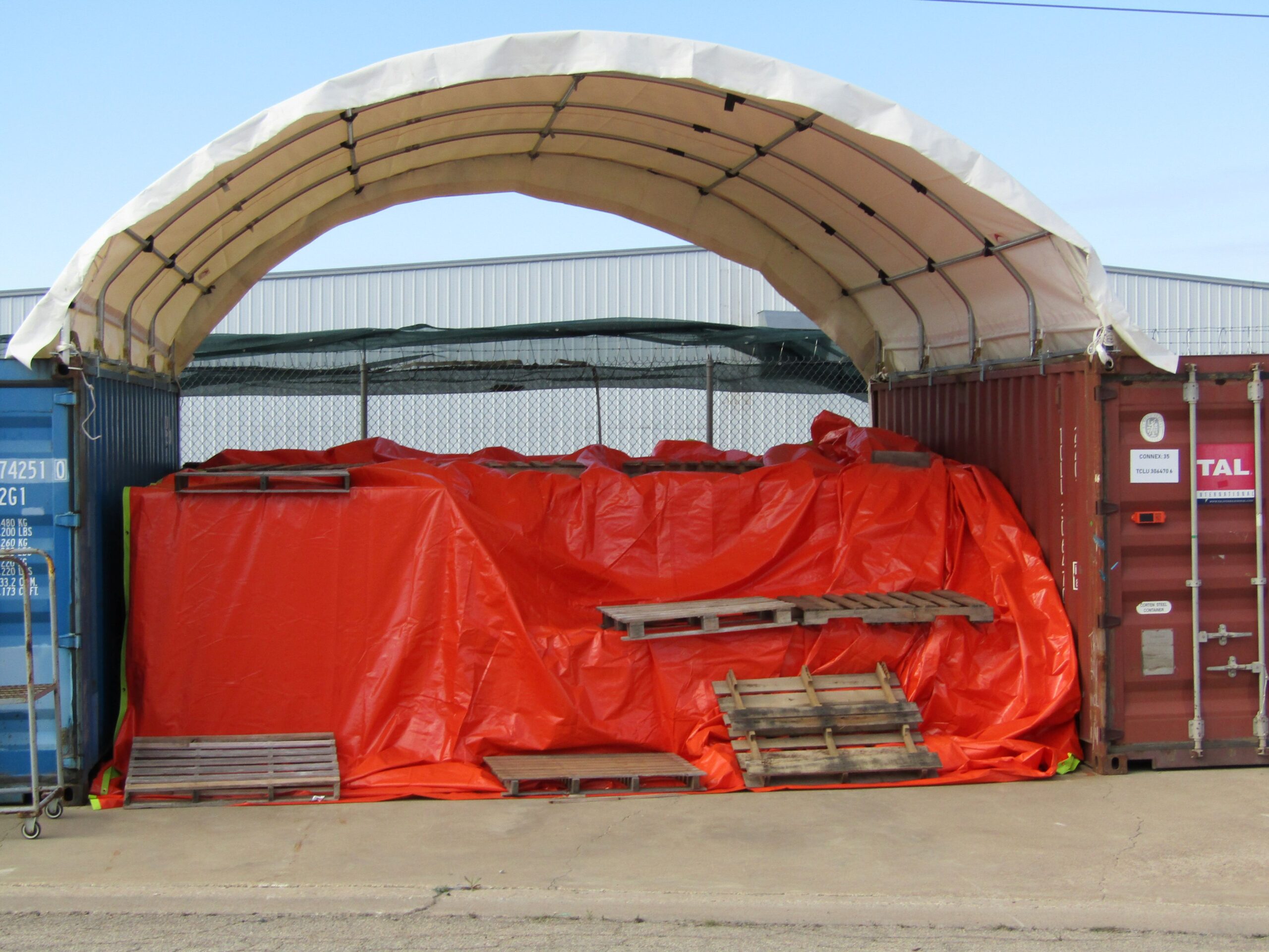 Container shelter arching between a blue and a rust-red shipping container over tarp-covered stored material