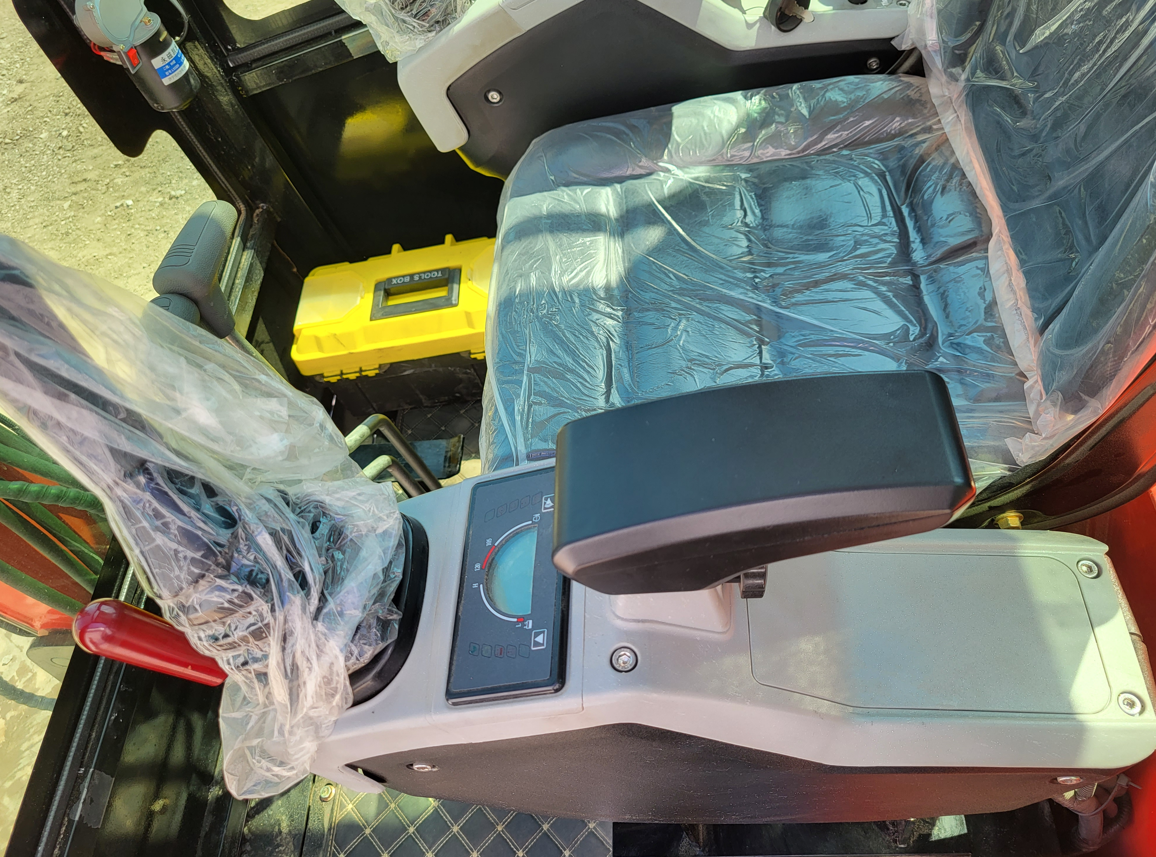 Mini excavator cab interior: operator seat and joysticks in factory wrap, gauge display, tool box on the floor