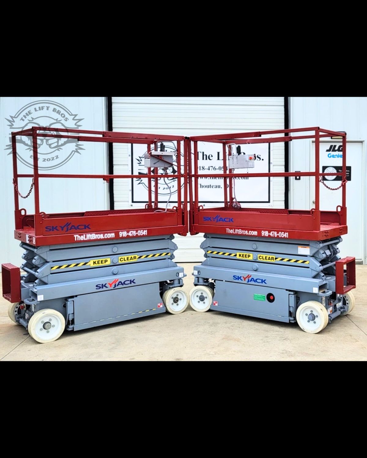 Two Skyjack scissor lifts side by side, lowered, red guardrail platforms on gray chassis with non-marking tires