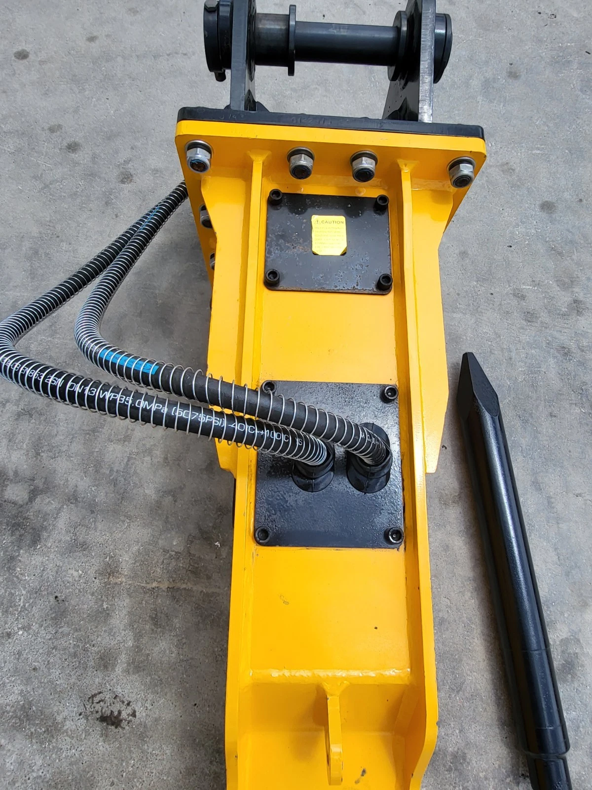 Top of the yellow TOFT750 breaker: bolted valve covers, caution decal, hydraulic hoses and a spare chisel