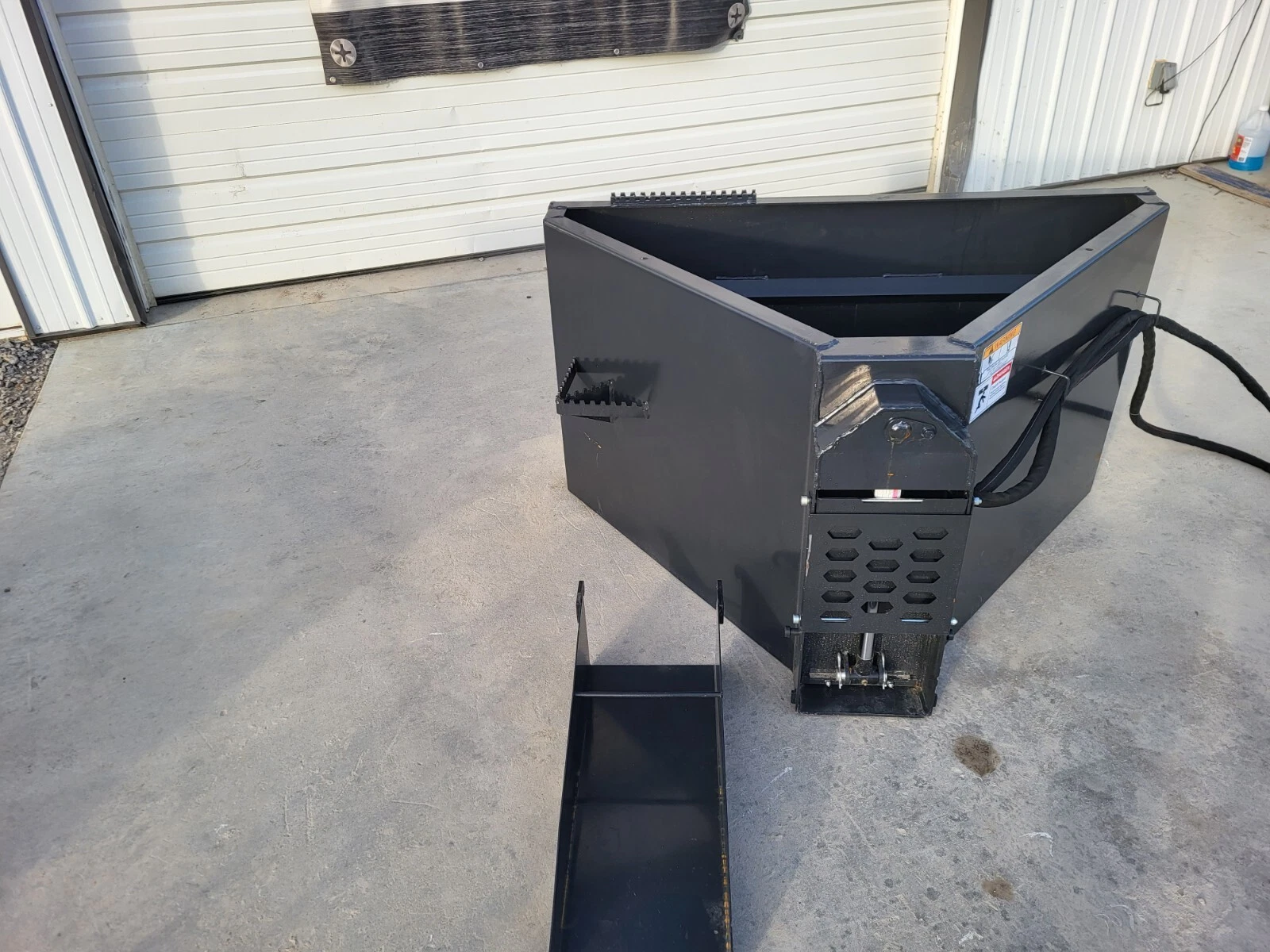 Concrete bucket with gate housing and hydraulic hose coiled at the side, pour chute on the ground