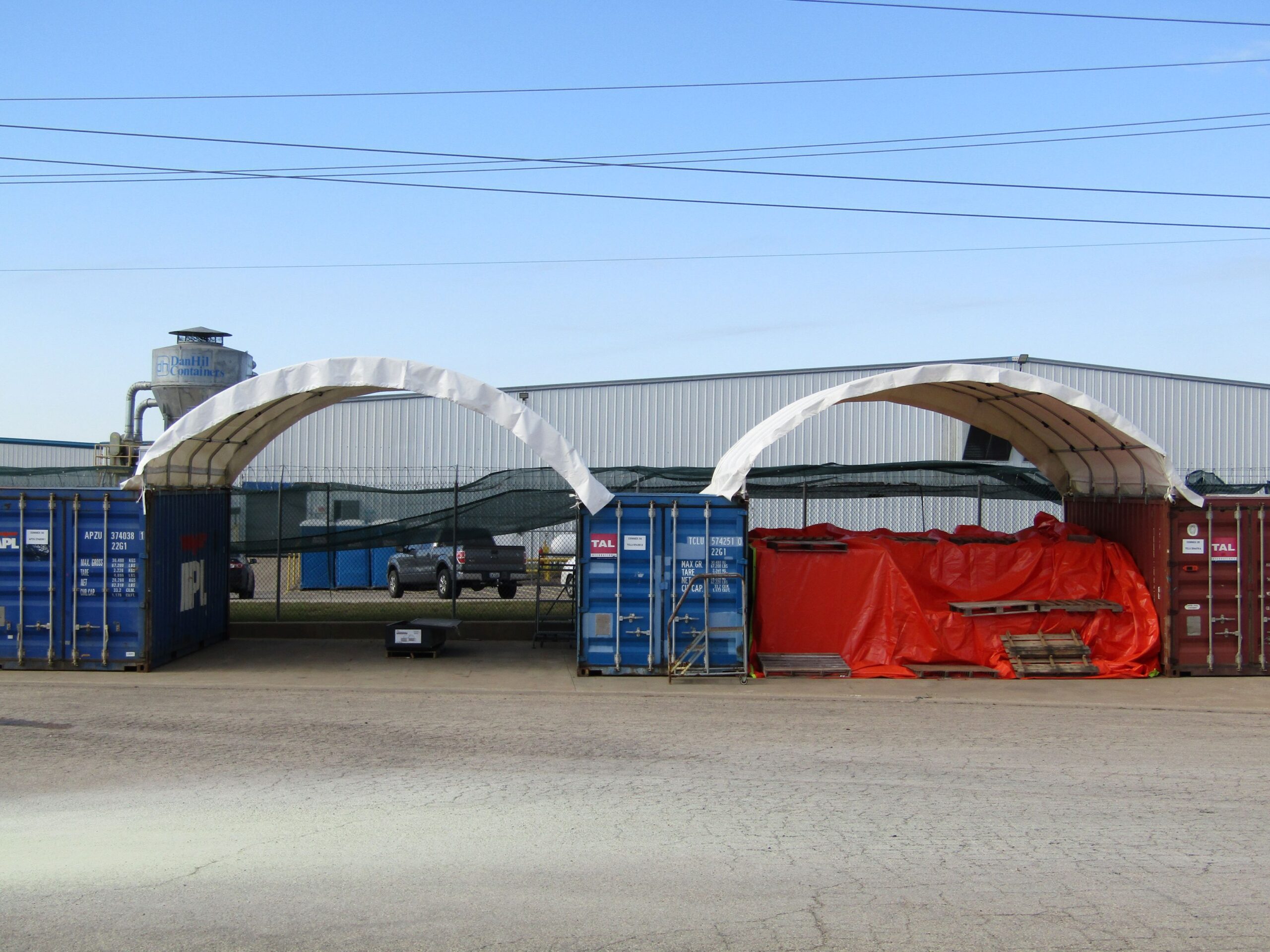 Two white container shelters installed across shipping containers, sheltering stored pallets and equipment