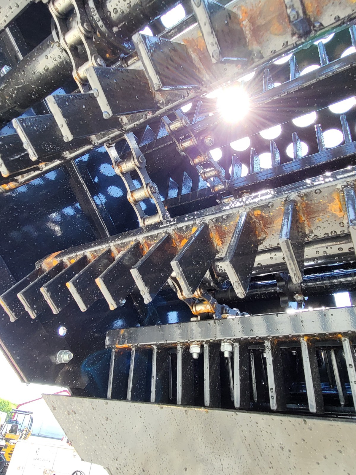 Underside of the landscape rake: drive chains and rows of angled steel scoop teeth on the drum, perforated grate above