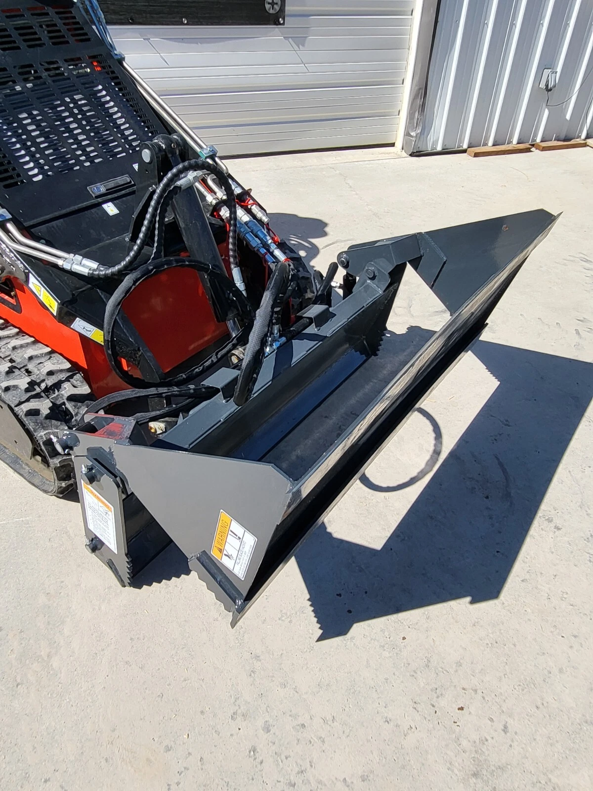 4-in-1 bucket held level on an orange mini skid steer, serrated jaw edge running the full width