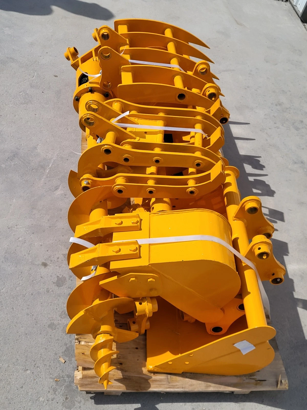 Yellow attachment package lengthwise on the pallet: rows of grapple and rake tines, buckets and auger bit