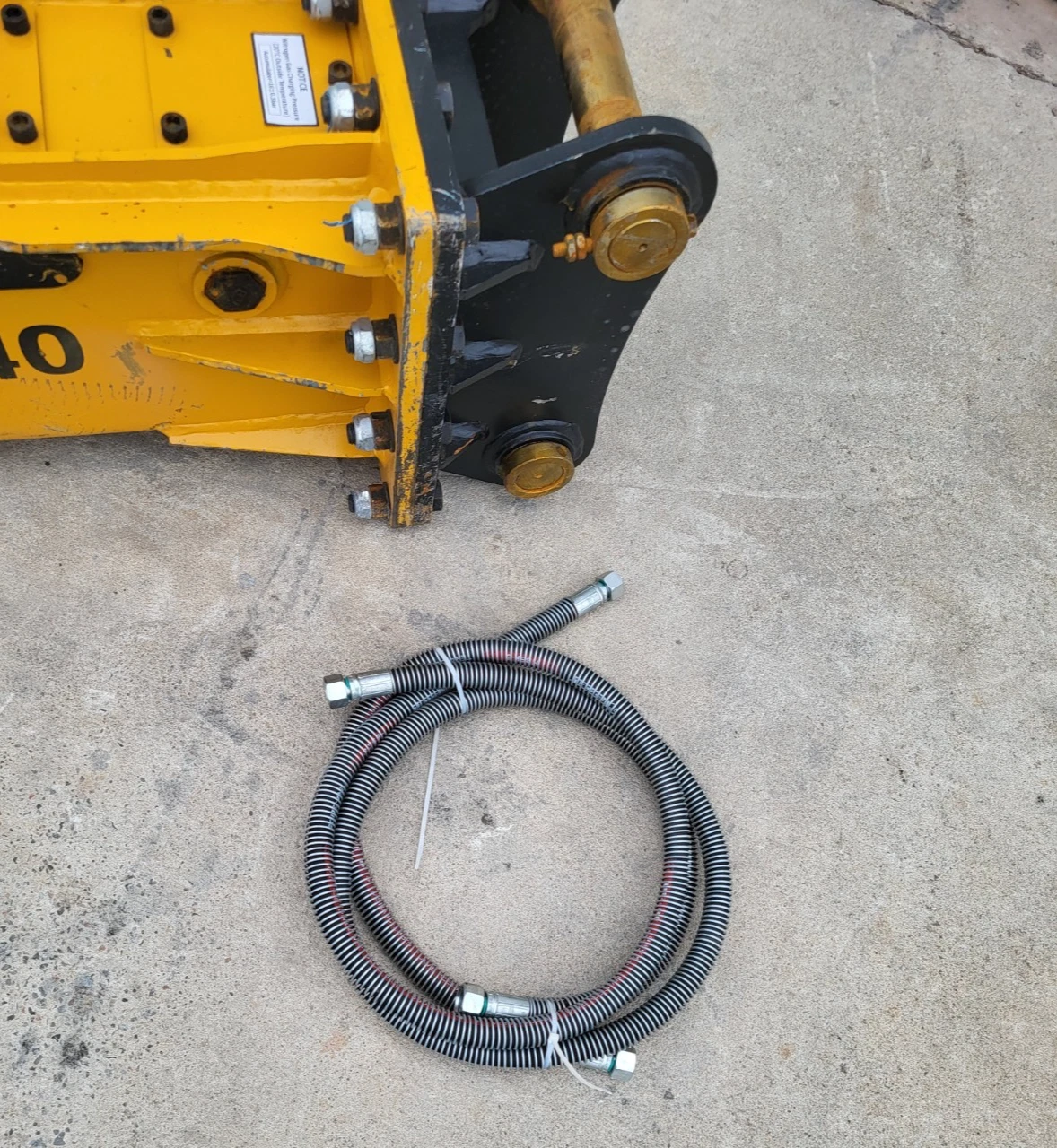Rear of the SB40 breaker with black mount and brass pins, coiled hydraulic hose pair alongside