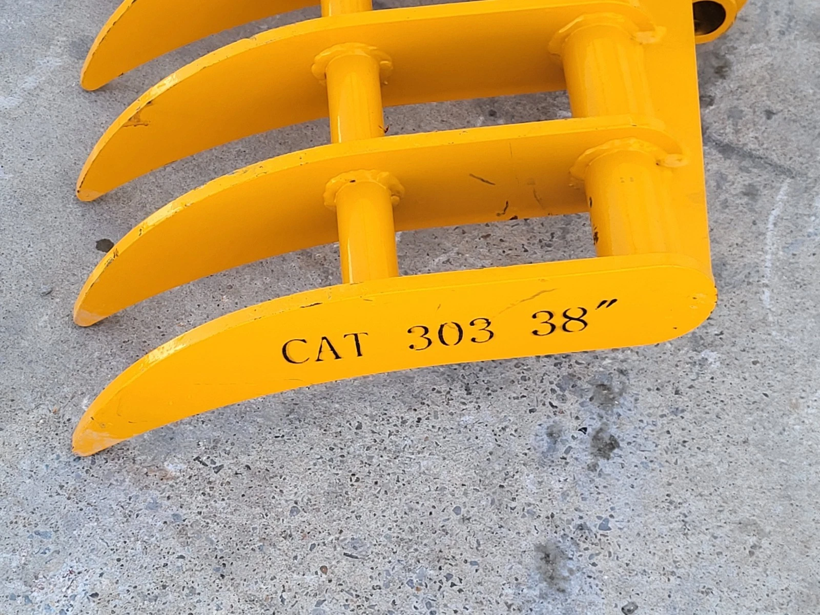 Yellow rake's outer tine bar stamped CAT 303 38 inch, tubular cross-braces above the curved tines