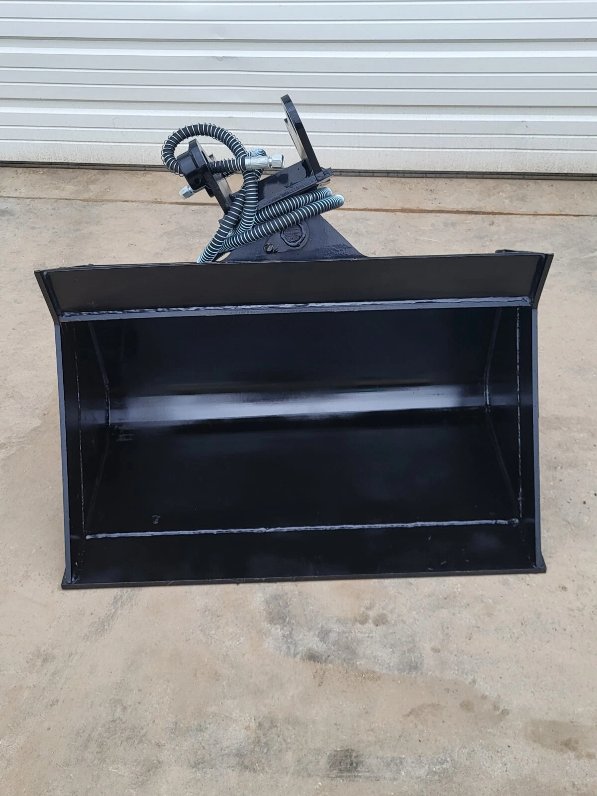 Black hydraulic tilt bucket from the front, deep open shell with tilt coupler and coiled hoses