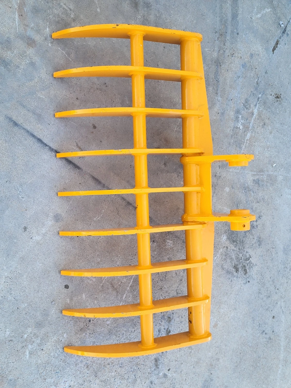 Yellow excavator rake from above, curved tines on a tubular frame with side-mounted pin bosses