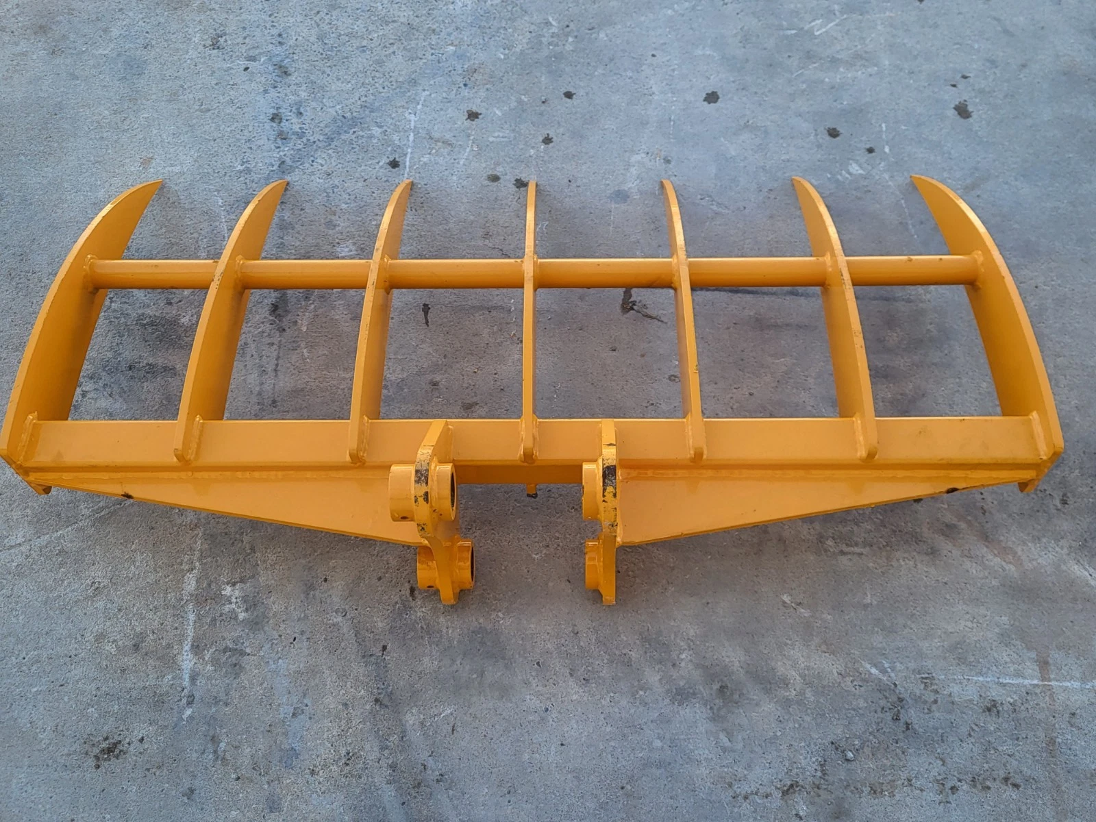 Yellow excavator rake from above, seven curved tines on a tubular frame with twin pin bosses centered