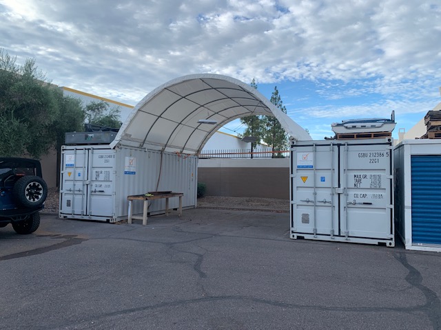 Container shelter with white PVC cover spanning two white shipping containers on galvanized arches