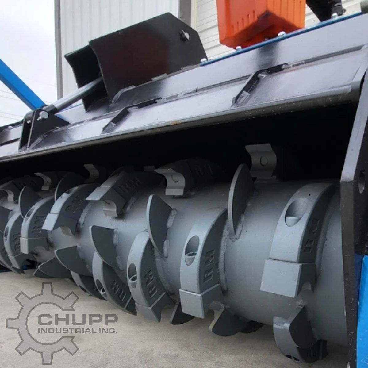 Drum mulcher rotor up close: spiral rows of bolt-on cutting teeth on the gray anti-wind drum under the deflector