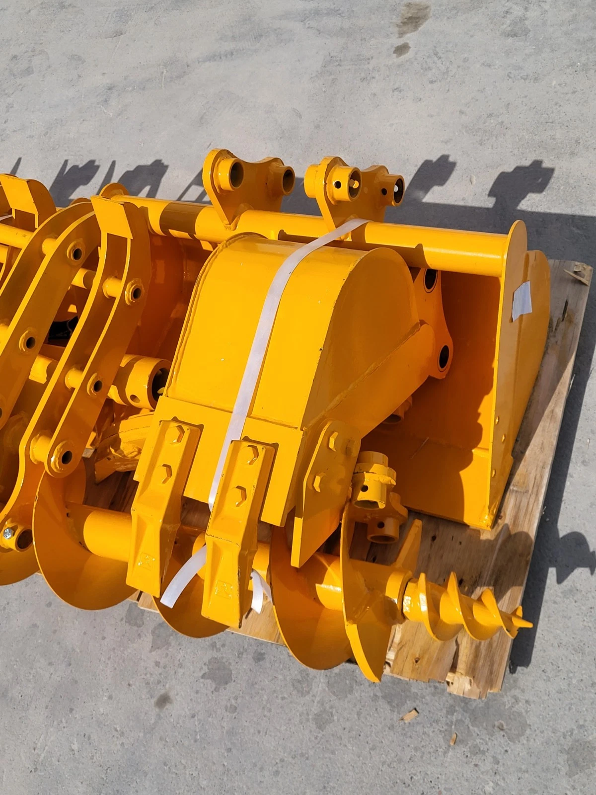 Yellow excavator bucket with twin pin lugs on the pallet, ripper shanks and auger bit alongside