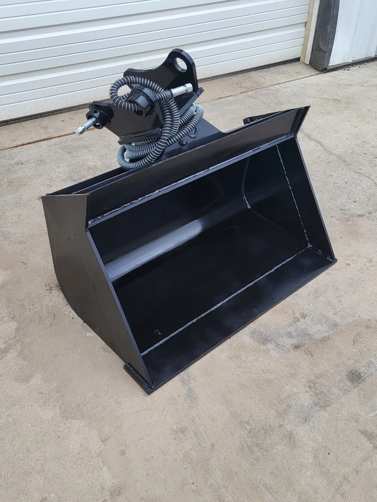 Black hydraulic tilt bucket tipped forward, tilt coupler with pin eye and coiled hydraulic hoses