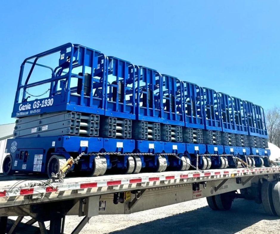 Row of blue Genie GS-1930 scissor lifts chained down side by side on a flatbed trailer