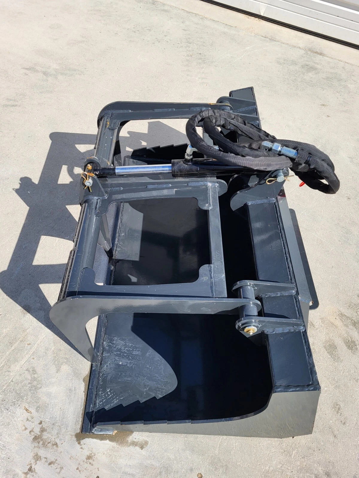 Gray flat-bottom grapple bucket, grapple arm open over the shell with hydraulic cylinder and hoses