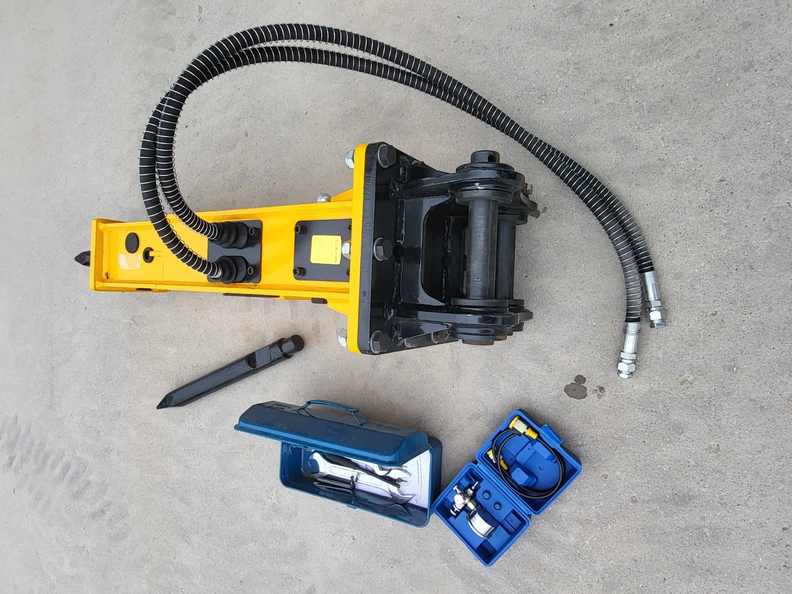Yellow breaker from the bracket end: black top-mount bracket and pin, coiled hoses, chisel and tool boxes