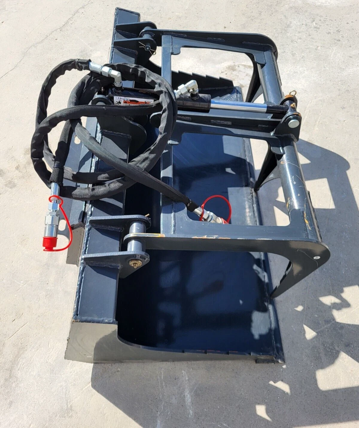 Gray flat-bottom grapple bucket with open grapple arm, hydraulic cylinder and red-capped coupler