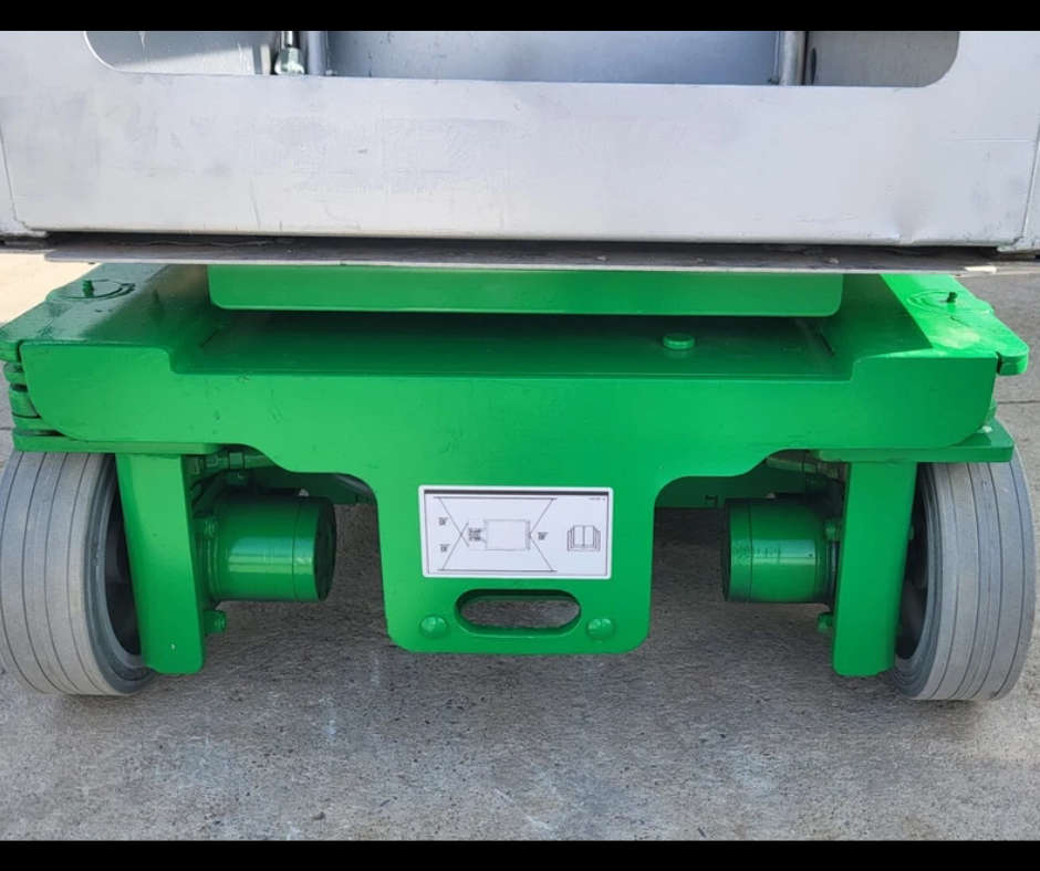 Green vertical mast lift base up close: solid non-marking tires, drive wheels and load decal under the platform