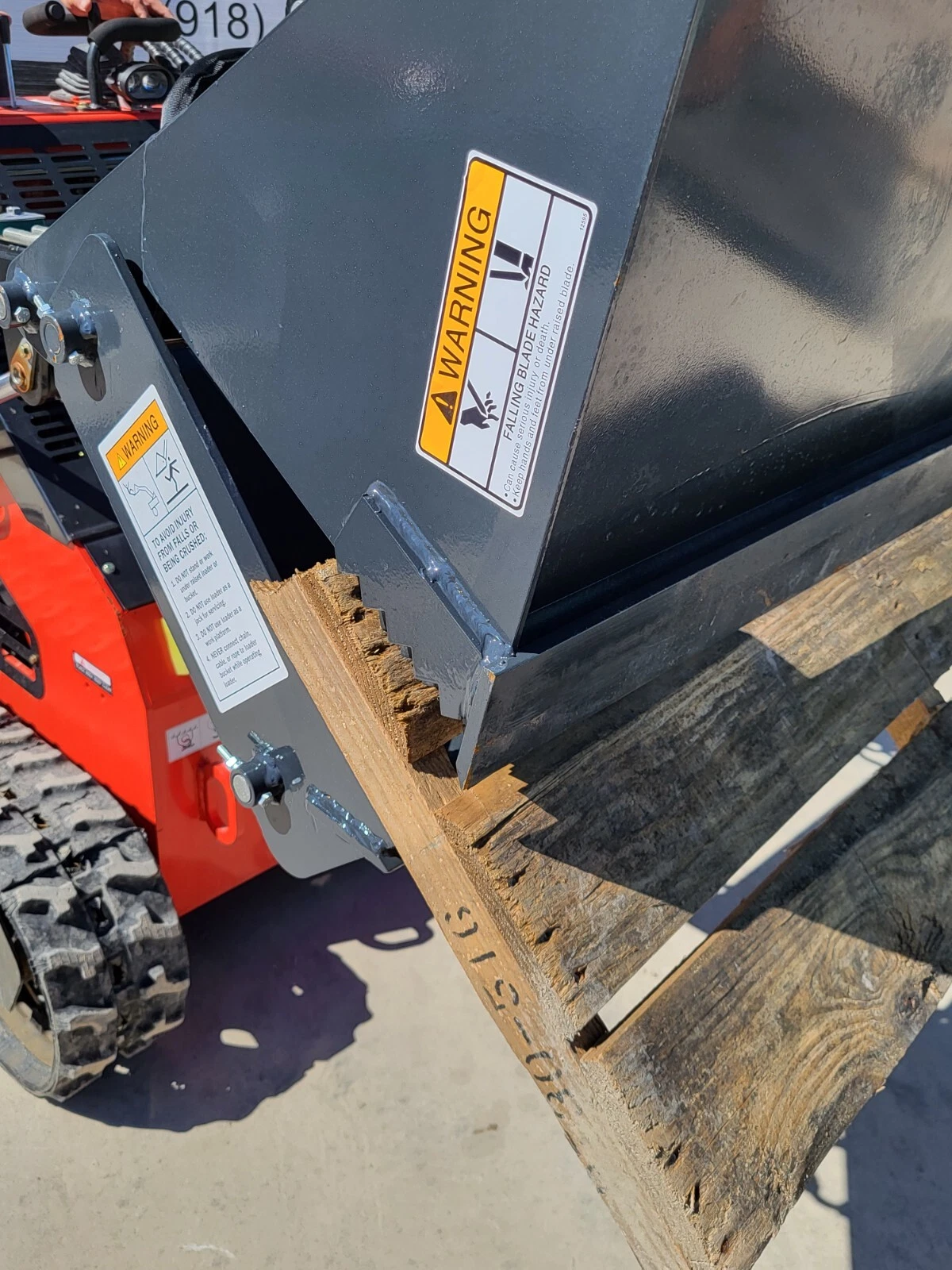 4-in-1 bucket's serrated clamshell jaw gripping a wooden pallet board, falling-blade warning decal on the shell