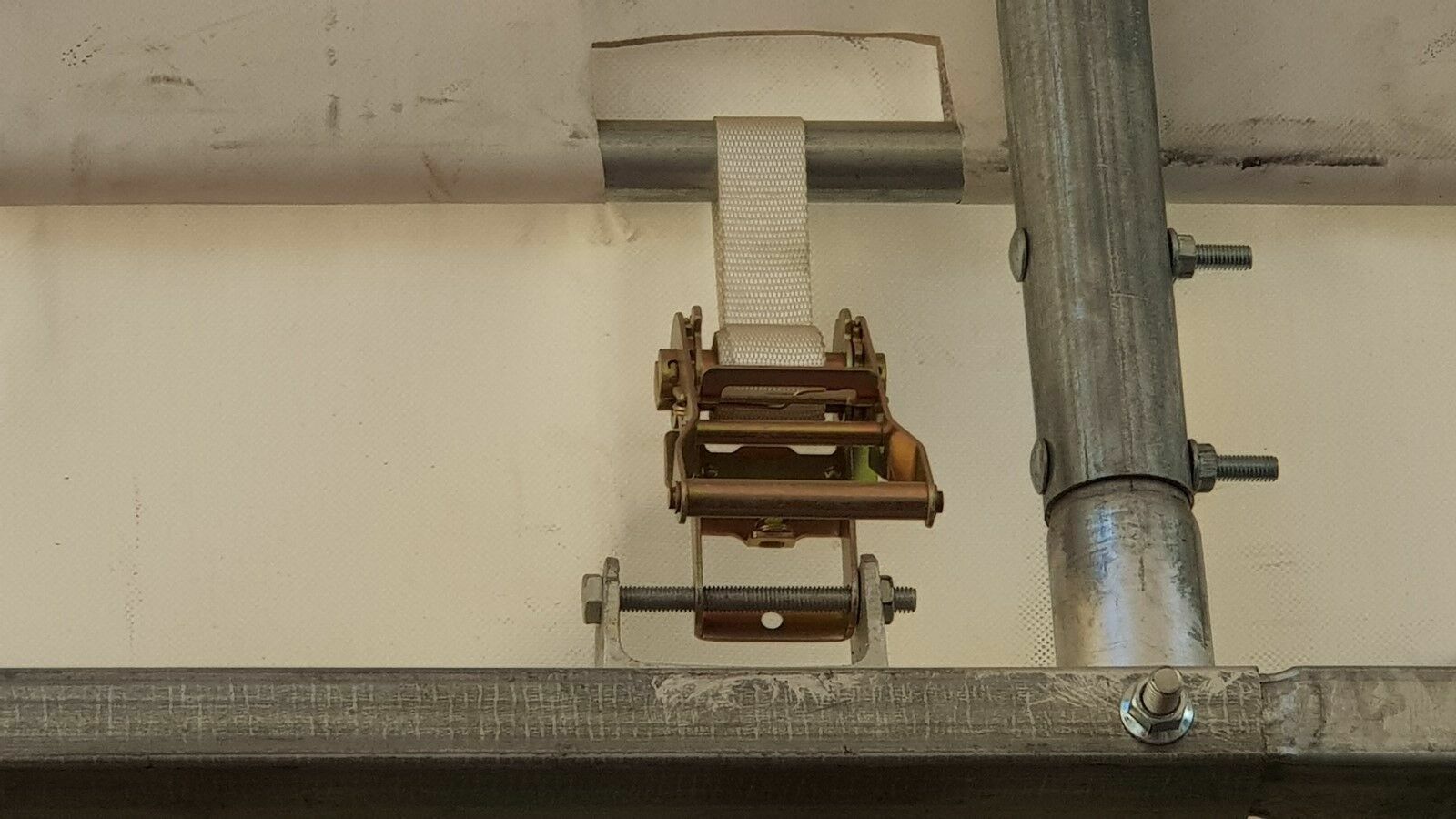 Ratchet strap tensioner clamped to a galvanized truss tube, tightening the white PVC shelter cover