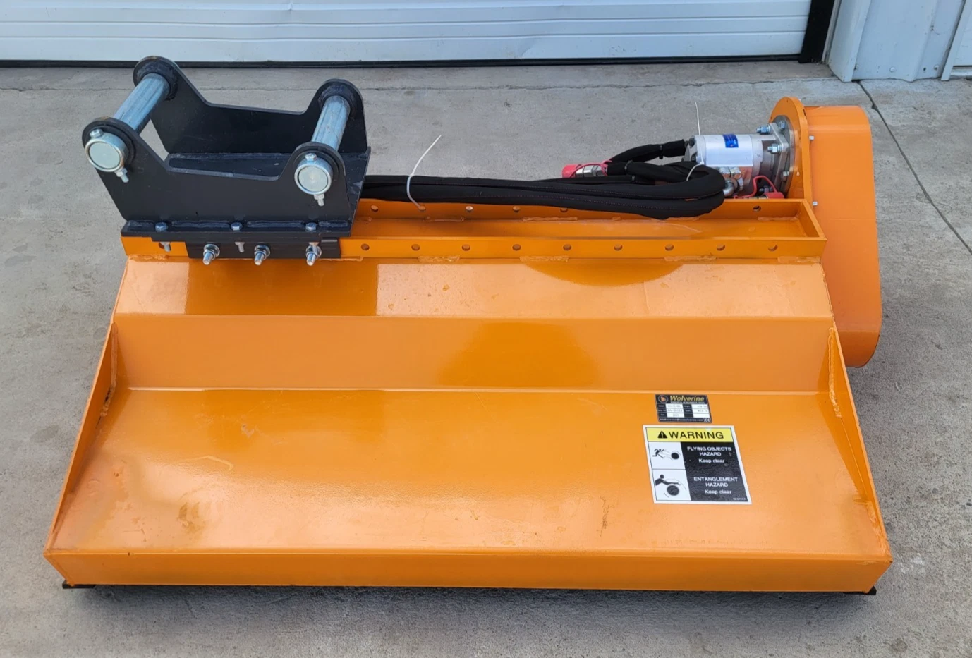 Orange flail mower with black excavator mount bracket, hydraulic motor and warning decal on the deck