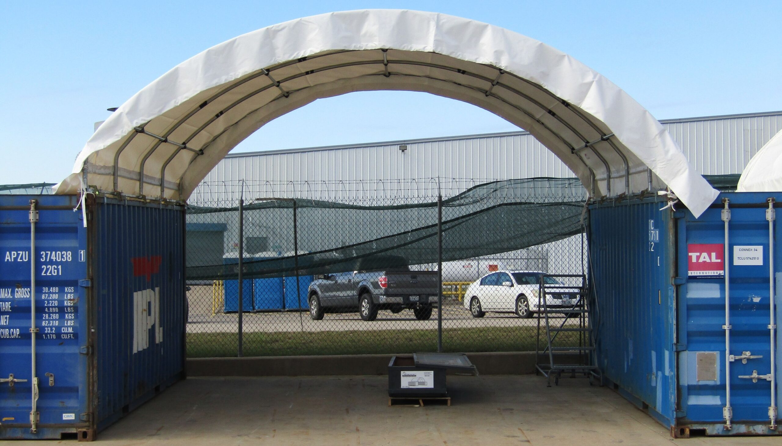 White container shelter forming an open-ended bay between two blue shipping containers