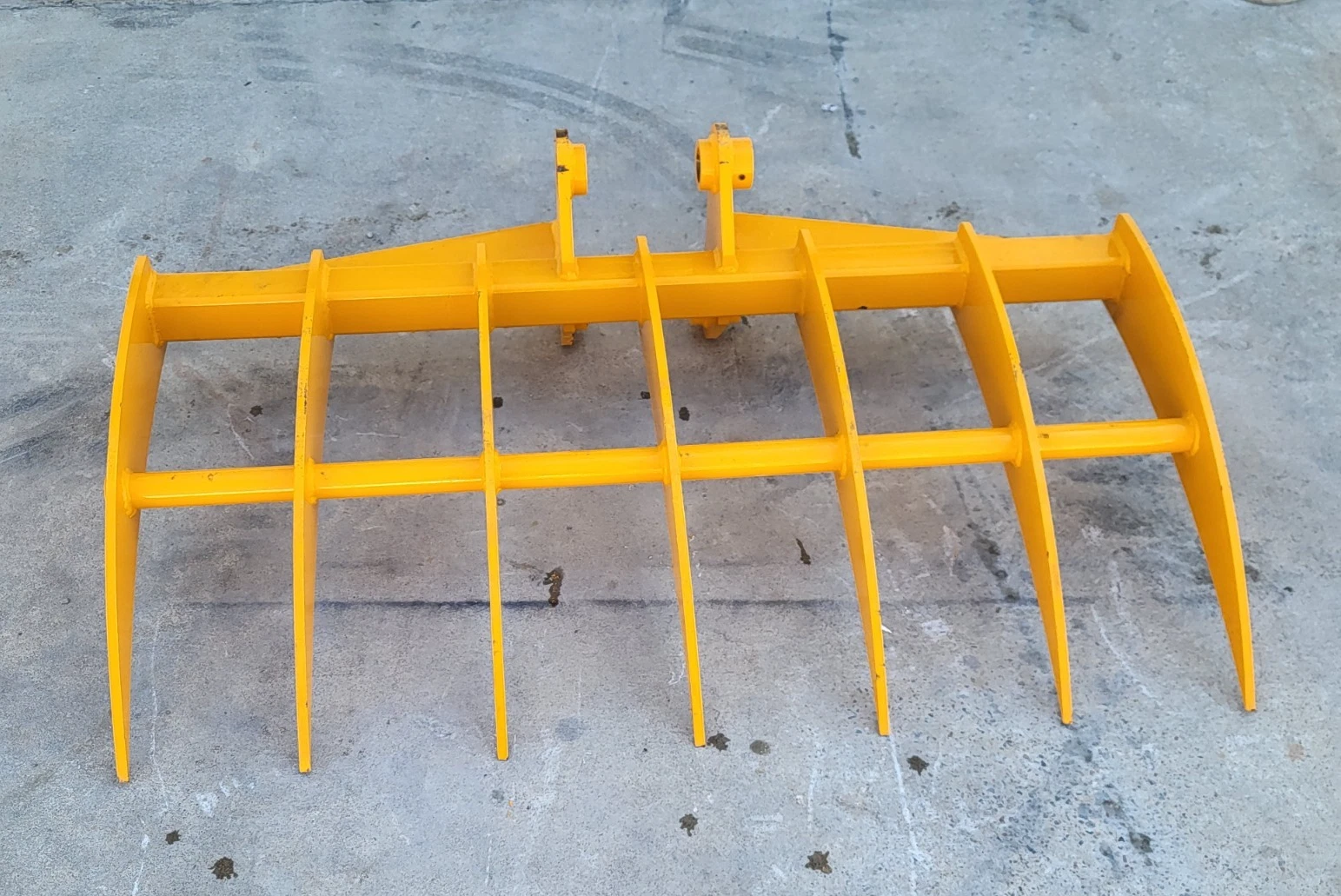 Yellow excavator rake seen from above, seven curved tines and twin pin-boss mounts on the frame