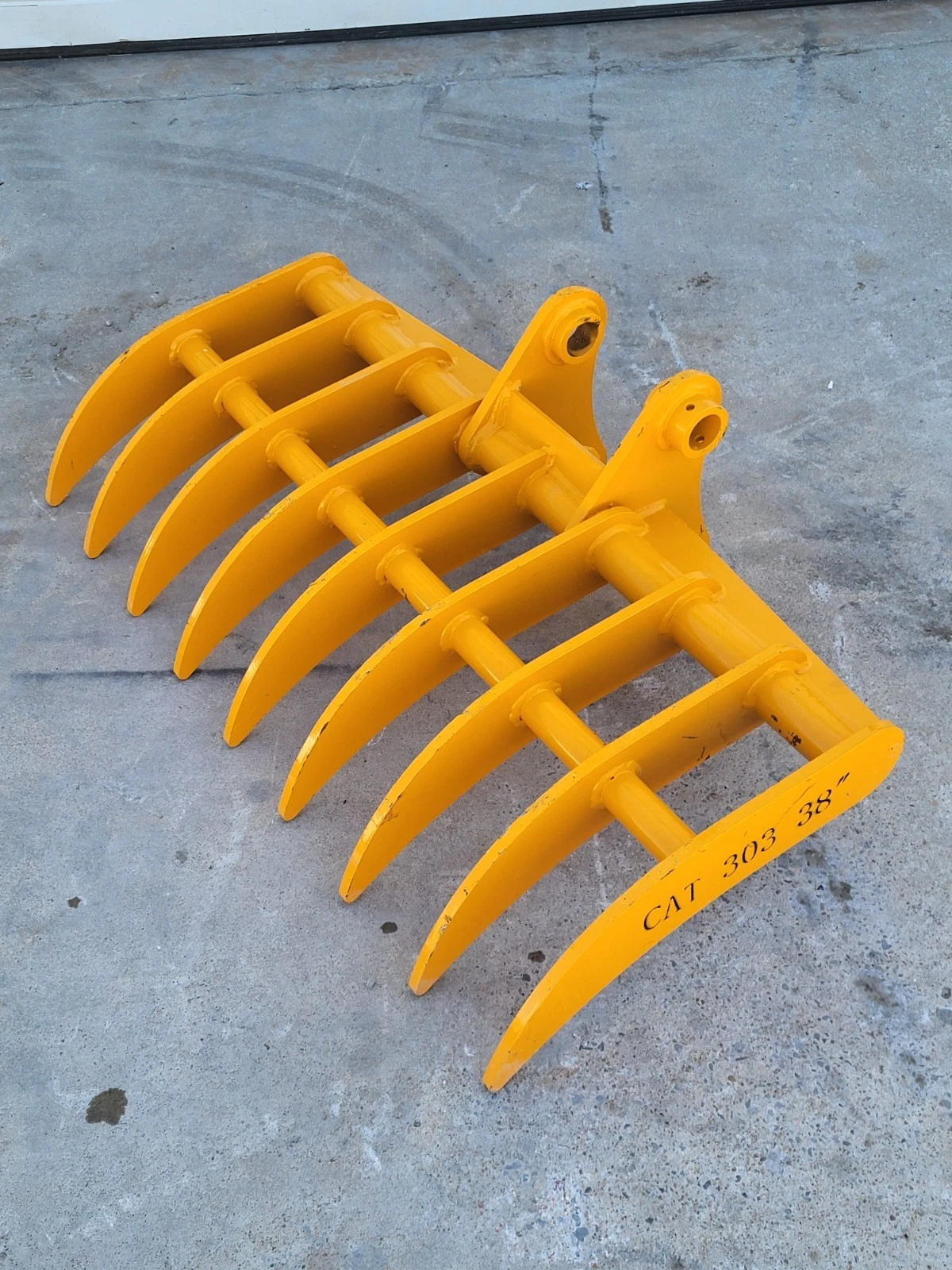 Yellow excavator root rake, eight curved tines on a tubular frame with twin pin-boss mounts