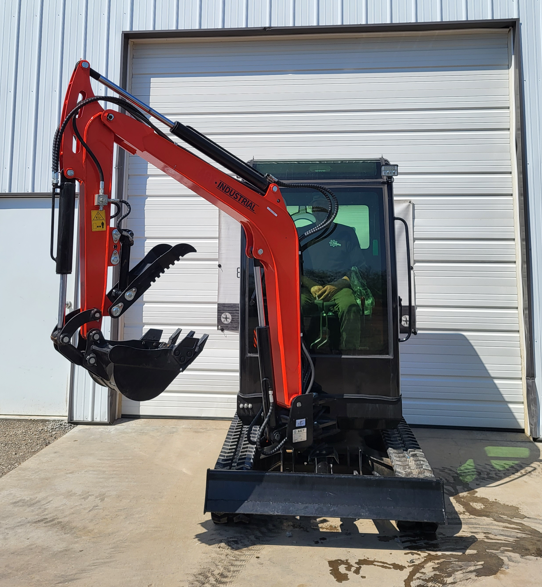 QNT50R mini excavator from the front: dozer blade down, rubber tracks, boom folded with thumb and toothed bucket