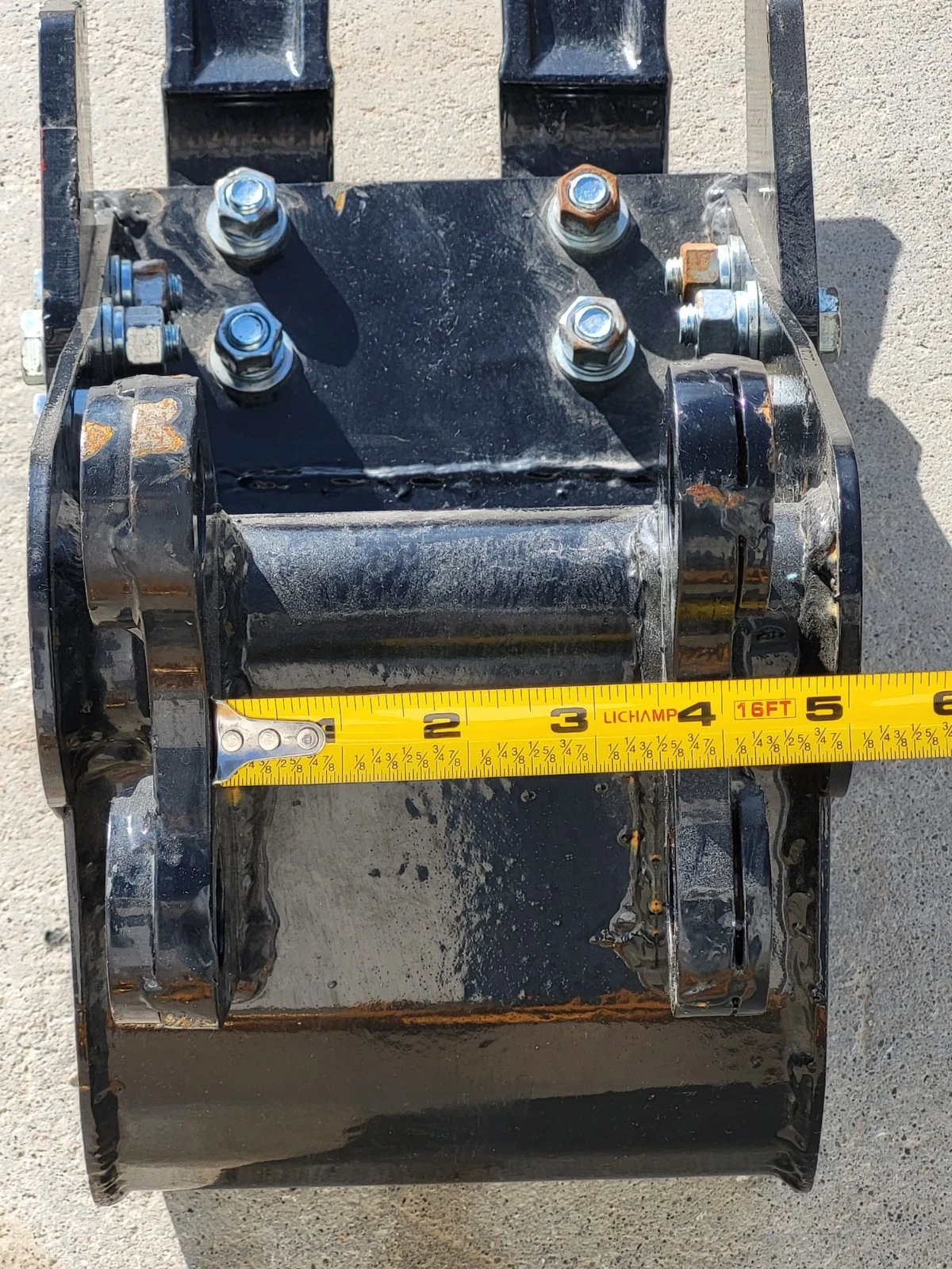 Tape measure across the trenching bucket's bolted hitch and pin bosses, past the four-inch mark