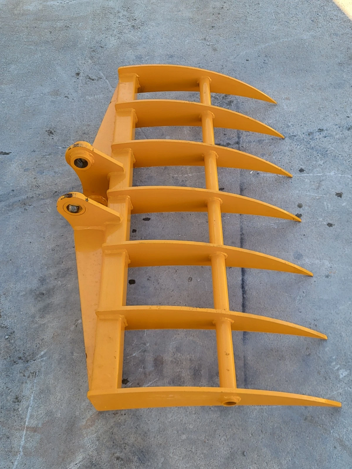 Yellow 46-inch excavator rake at an angle, curved tines fanned out and twin pin-boss mounting ears
