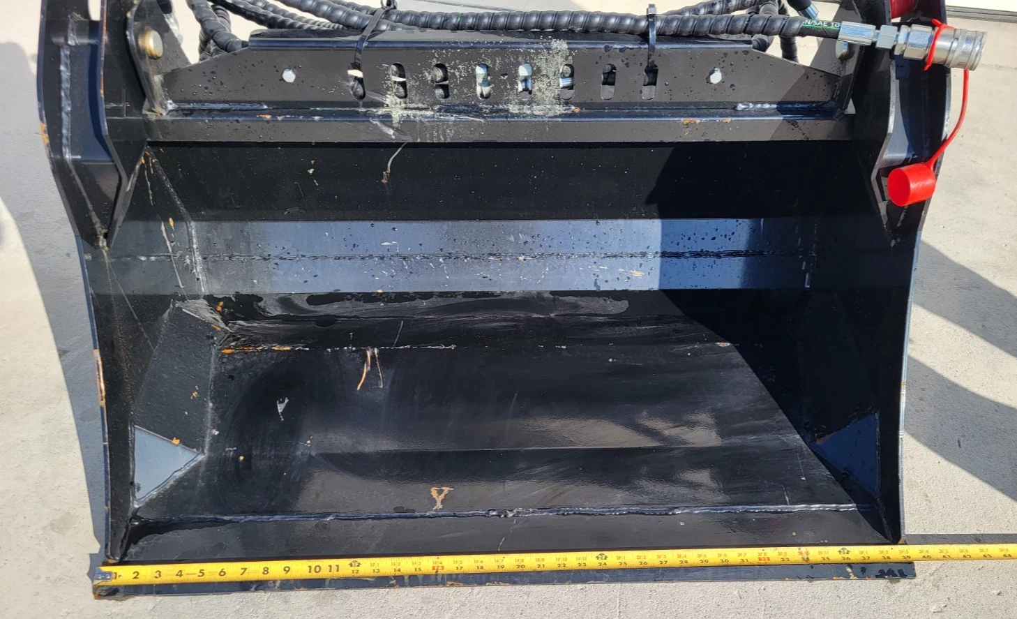 Black 4-in-1 bucket's open interior, hydraulic hoses and a red-capped coupler above, tape measure along the edge