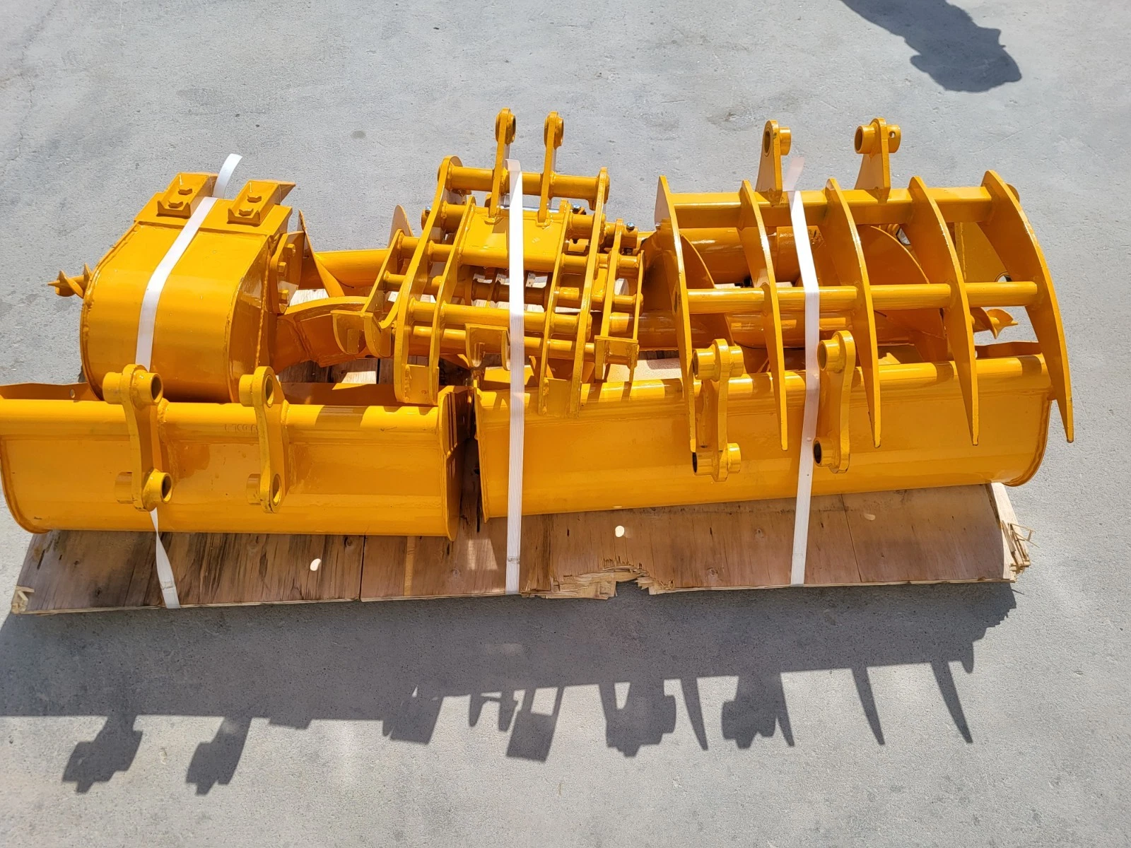 Yellow mini excavator attachment set banded to a pallet: buckets, grapple tines, rake and auger flighting