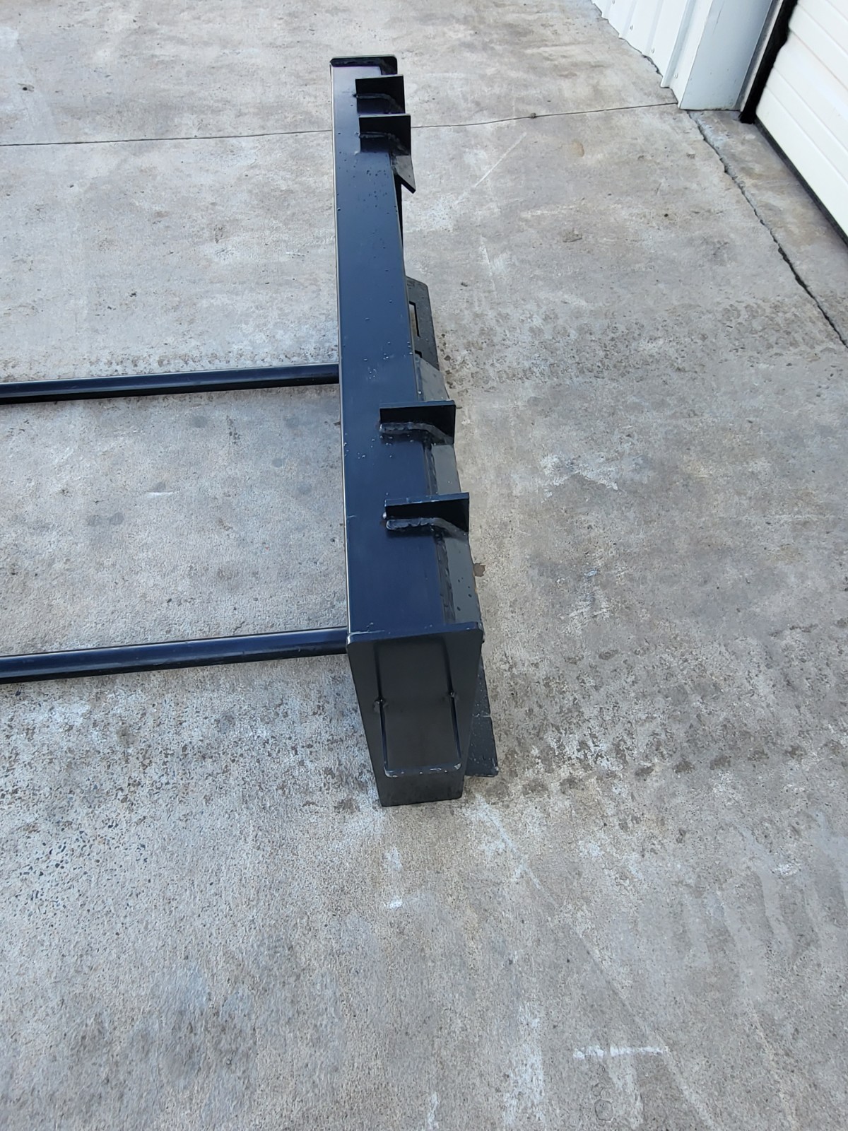 JCT double bale spear from the side: black quick-attach frame with the two round tine bars extending along the ground