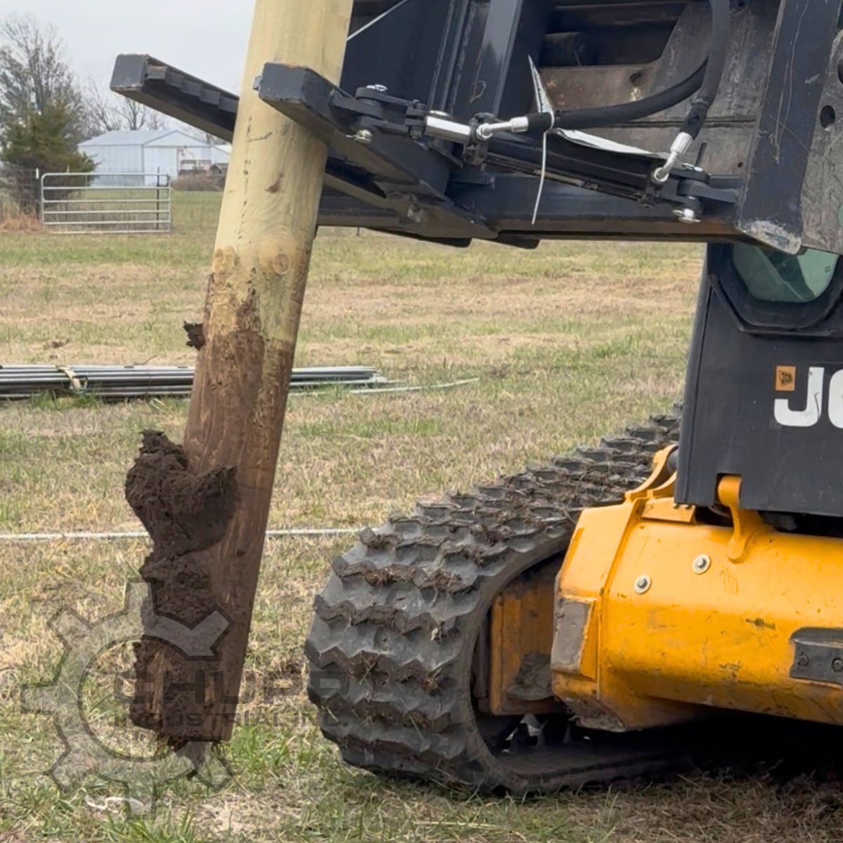 Tree and post puller on a JCB track loader, gripping a muddy wooden fence post pulled out of the ground