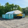 White arched shelter over two teal shipping containers with a yellow boom lift parked under the open end