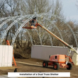 Boom lift raising galvanized dual-truss arches between two shipping containers during shelter installation