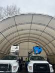 White PVC container shelter from inside: arched steel dual trusses over two bucket trucks parked under the canopy