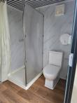 Portable restroom interior: glass shower stall with curtain, ceramic toilet, wall vent fan and paper dispenser