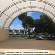 Inside a white dual-truss shelter beside a container wall, open end showing palm trees and a waterway