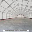 Inside a white dual truss fabric building, galvanized trusses overhead and a concrete floor, end wall in the distance