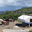 Distant white shelter tent beside stacked shipping containers and pipe, tropical hillside and coast