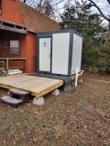 White hard-sided portable restroom unit, dark charcoal corner frame, single door, PVC drain lines connected at the base