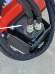 Hydraulic motor on the power rake, two sleeved hoses bolted to its ports on the black housing above the red frame