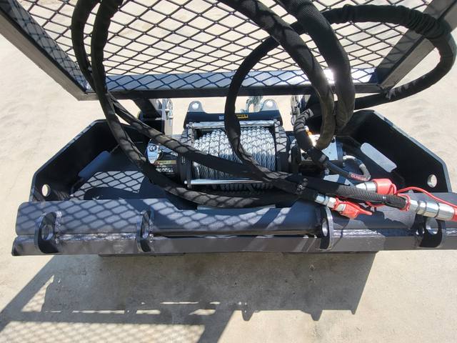 Forestry winch in its frame: cable drum, hydraulic motor, hoses and red-capped couplers under a mesh deck