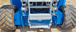 Blue scissor lift rear undercarriage: knobby rough-terrain tires, gray scissor-arm pivots and fold-down steel access steps