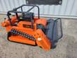 Orange tracked flail mower from the front corner: gas engine, roll cage, flail head with mesh guard
