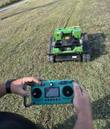 Green tracked remote-control mower on a grass slope, operator holding the teal handheld remote in the foreground